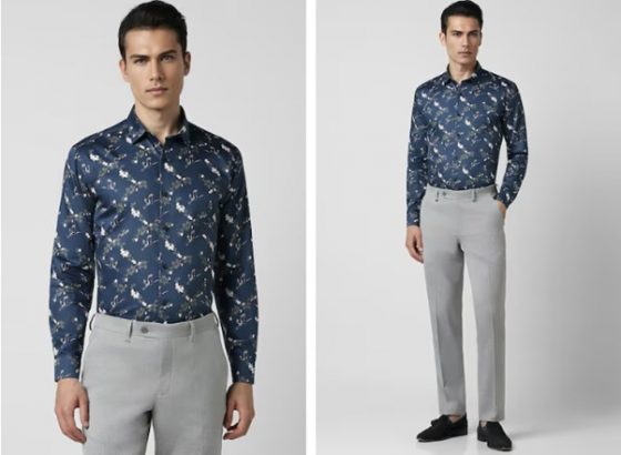 Man in a blue patterned shirt and light gray trousers, standing against a white background.